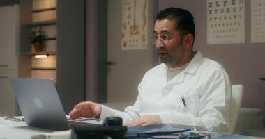 A man in a white lab coat is sitting at a desk with a laptop video