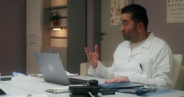 A man in a white lab coat is sitting at a desk with a laptop video