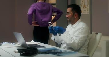 A man in a lab coat is sitting at a desk with a laptop video