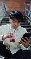 A man sitting on steps holding a drink and looking at his phone video