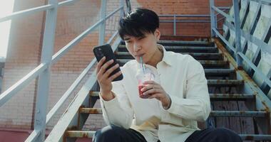 A man sitting on stairs holding a drink and looking at his phone video