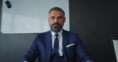 chat, front camera. A middle-aged man of European appearance, dressed in a business suit, looks directly at the camera, talking on a call in the office video