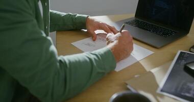 A young man of European appearance makes corrections in a drawing, sitting at a work table in front of an open but turned off laptop, close-up male hands, no face is visible video