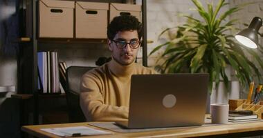 jovem encaracolado cabelos morena homem é interrompido a partir de trabalhando em uma computador portátil, leva fora dele óculos e parece em linha reta para dentro a Câmera com uma sério Veja enquanto sentado às uma mesa dentro a escritório video