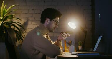 Young man in glasses with stubble on his face talking hands-free on a mobile phone while sitting at a table in front of an open laptop in the office, under the light of a lamp video