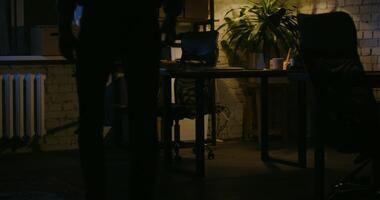 A young man with glasses, dressed in casual clothes, comes to the office at night, opens a laptop and starts working while sitting at a table under the light of a lamp video