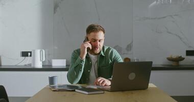 A young respectable man of European appearance is talking on a mobile phone, sitting at the kitchen table, with a laptop on it and a lot of work papers around video