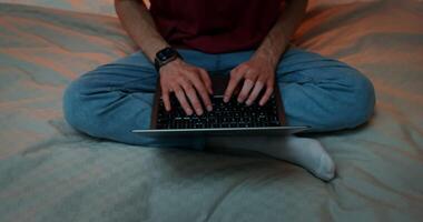 A man is typing on a laptop, standing on his feet, sitting in a lotus position on the bed, Close-up of male hands on the keyboard, no face, front view from above video