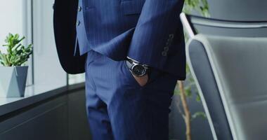 A respectable man of European appearance in a business suit, pulls out his hand from his trouser pocket while standing near the window in the office, close-up, the man's face is not visible video