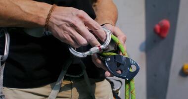 A European-looking man inserts a rope into a carabiner and attaches it to a safety harness, standing in front of a climbing wall, getting ready to go mountaineering in an indoor gym, close-up video