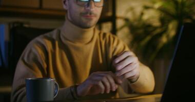 Young man in glasses with bristles on his face drinks coffee while sitting at a table in front of an open laptop and looks at the screen, shooting with closer to the face video