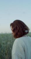 A young red-haired woman with a short haircut walks through the field among tall ears of wheat with a thoughtful look sorting through the ears. 4k, vertical , red komodo video