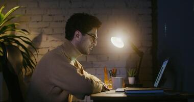 A young man wearing glasses with a stubble on his face is talking on a link, sitting at a table in front of an open laptop in the semi-darkness by the light of a lamp. shot from the side video