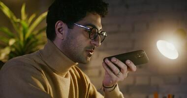 Young man in glasses with stubble on his face finishes talking on a speakerphone on a mobile phone while sitting at a table in an office under the light of a lamp, close-up video