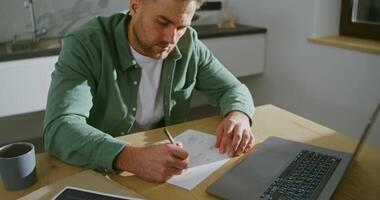 A young stylishly dressed man of European appearance makes corrections in the drawing, sitting at the kitchen table in front of an open laptop. Remote work of a designer from home video
