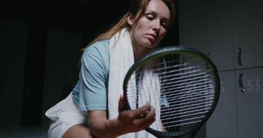Young beautiful woman tennis player is resting in the locker room after the match, sitting on a bench with a towel around her neck and twirling a tennis racket in her hands, close-up video
