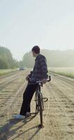 A young man with a camera around his neck stops on a gravel road near a field after cycling and admires the beauty of the surrounding fields. 4k vertical , red komodo video