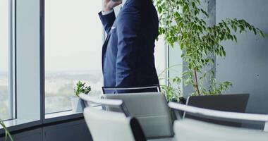 Respectable middle-aged man of European appearance with a gray beard, dressed in a business suit, talking on a mobile phone in a modern stylish office and looking out the window video