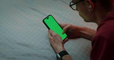 A young man uses a smartphone while lying on the bed. Green screen, copy space, top view video