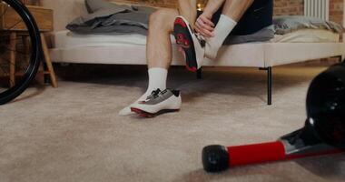 A man puts on cycling shoes for training on a cycling simulator at home in a stylish modern bedroom. Cardio workout at home. Indoor cycling video