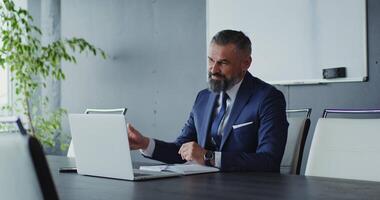 A respectable man of European appearance with a gray beard, dressed in a business suit, talks calling on a laptop and drinks coffee, sitting at a table in the office video