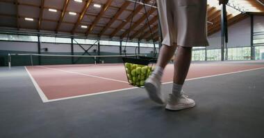 A man carries a basket full of tennis balls in one hand and a tennis racket in the other hand, walking on an indoor tennis court, preparing for a workout, the man's face is not visible video