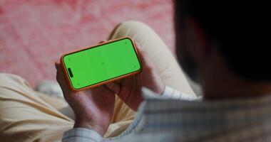 A man in casual clothes holds a mobile phone with a green screen turned horizontally while watching a indoors. Top view, no face, close-up of a man's hands video