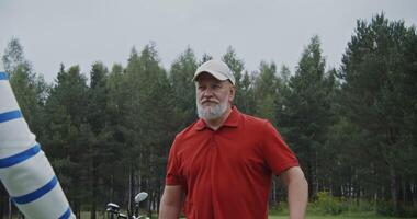 un personnes âgées aux cheveux gris homme avec une barbe dans une base-ball casquette, habillé dans élégant tenue de sport, pourparlers à un autre homme tandis que permanent sur une le golf cours. Extérieur tournage dans lumière du jour video