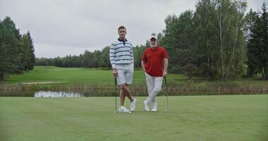 Two men of different ages are standing on the golf course with their legs crossed and leaning on golf clubs and smiling while looking at the camera. Father and son spend time playing golf together video