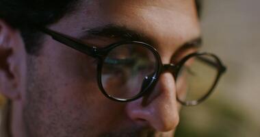A young brunette man in glasses with stubble on his face looks intently at the laptop screen while typing a text, a close-up of the face video