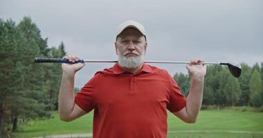 An elderly man of European appearance with a gray beard and mustache is looking seriously at the camera while standing on a golf course, and holding a golf club with both hands behind his head video