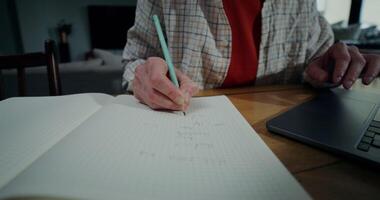 A woman in casual clothes writes in a notebook and uses a laptop while sitting at a table indoors, Close-up of women's hands, no face video