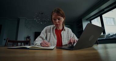A woman makes notes in a notebook and types on a laptop, sitting at a table in a home interior with a serious look video