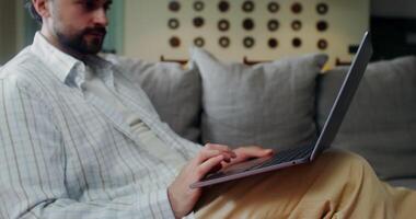 A stylish man in casual clothes starts a call on a laptop smiling and waving hello while sitting on a sofa at home video