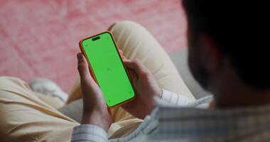 A man in casual clothes holds a mobile phone with green screen in his hand, sitting on the couch at home. Top view, no face, close-up of a man's hands video