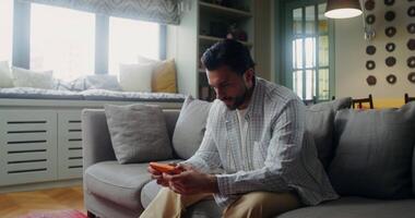 A man in casual clothes smiles while playing a game on a mobile phone while sitting on the couch at home video
