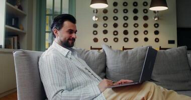 A stylish man in casual clothes starts a call on a laptop smiling, and waving hello while sitting on a sofa in a stylish living room video