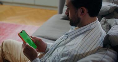 A bearded man in casual clothes uses a mobile phone with a green screen, scrolling the screen with his finger while sitting on a sofa indoors video