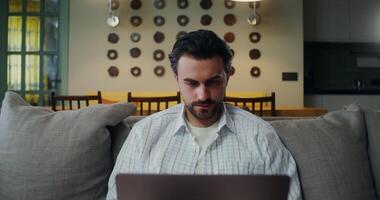 A young man in casual clothes is interrupted from using a laptop and looks at the camera, with a smile while sitting on the couch at home video