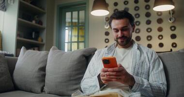 A man in casual clothes laughs when he uses a mobile phone while sitting on the couch at home video