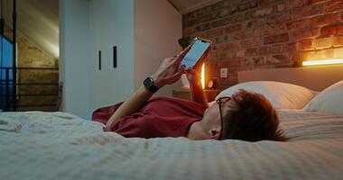 A young man in glasses dressed in casual clothes is typing text on a mobile phone, while lying across the bed in the evening in a stylish home interior video