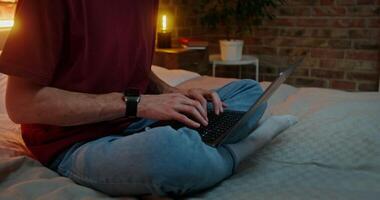 A young man in glasses is typing on a laptop while sitting on a bed in a lotus position in a stylish home interior in the evening. moves from typing hands to man's face video