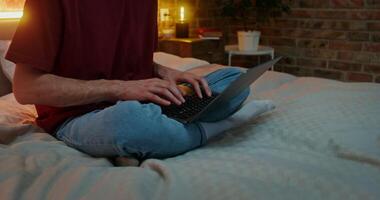 A young man in casual clothes is typing on a laptop while sitting on a bed in the light of lamps. Close-up of male hands and laptop, the man's face is not visible video