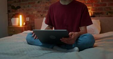 A young man with headphones in his ears is talking on a chat on a laptop while sitting on a bed in a lotus position. in the evening, the movement of the laptop to the man's face video