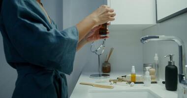 A young woman in a bathrobe, with a towel around her head, applies a moisturizer with a pipette to her face and rubs it with her hands, looking into the mirror in the bathroom video