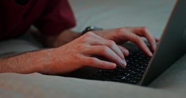 A young man with headphones in his ears is typing on a laptop while lying on the bed, the moves from the laptop keyboard to the man's face video