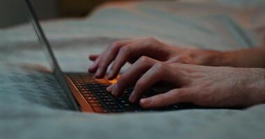 A man is typing on a laptop keyboard while lying on a bed in the evening, close-up of male hands, no face, unrecognizable identity video