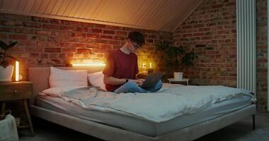 A young man in glasses with an earring in his ear is typing on a laptop, while sitting on a bed in a lotus position in a stylish home interior in the evening video