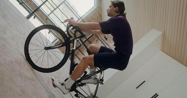 Young focused man rides a bike simulator in a modern home interior. Vertical filming transforms into horizontal, circular camera movement video