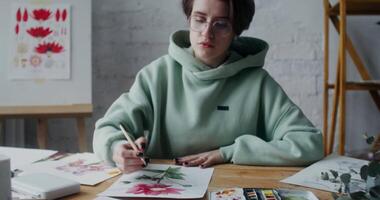 A young woman picks up watercolor paint from a palette and draws flowers on paper in an art workshop, working in daylight video